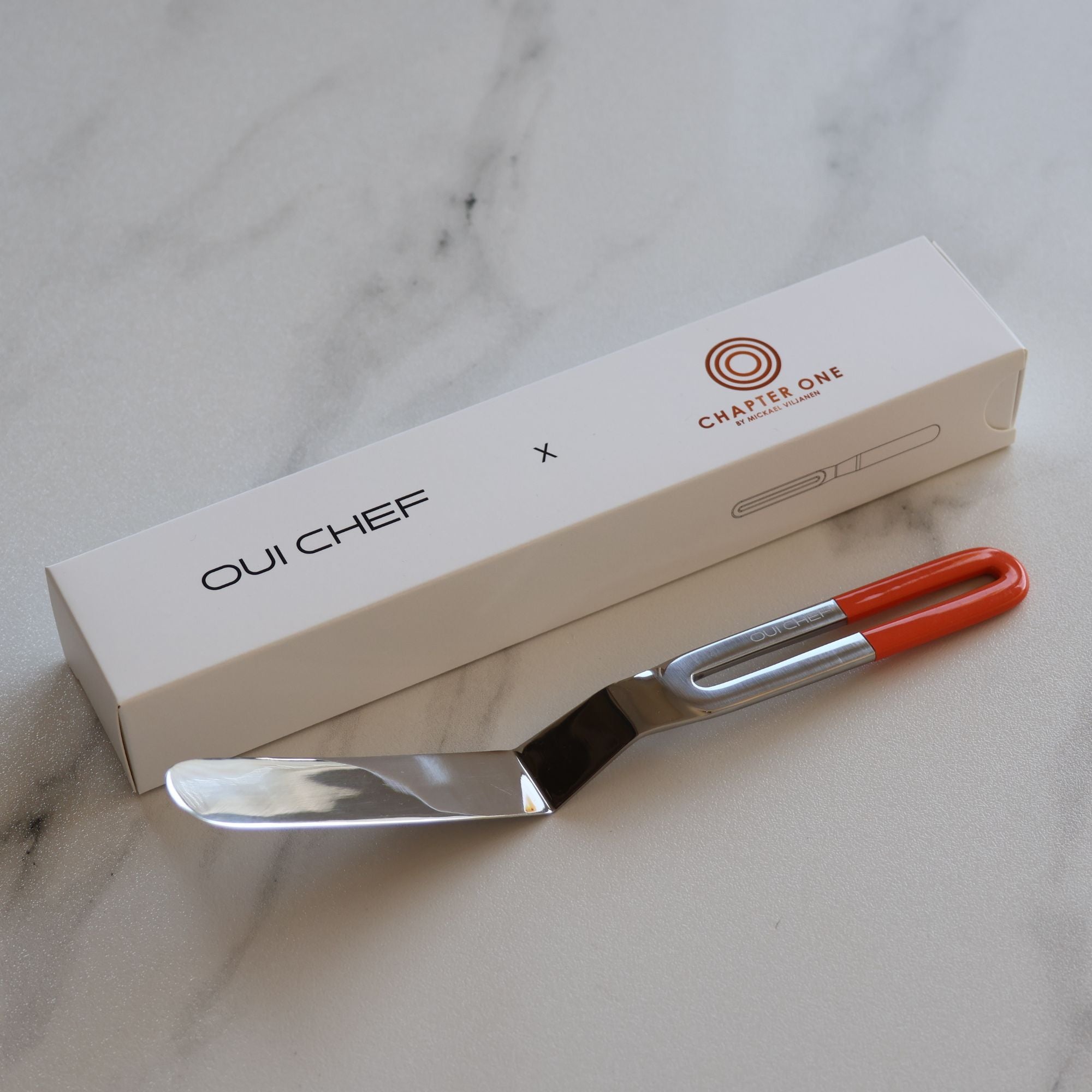 Stainless steel palette knife with an edge-to-edge sharp blade and powdered orange handle sitting on a marble countertop beside custom Chapter One box packaging.