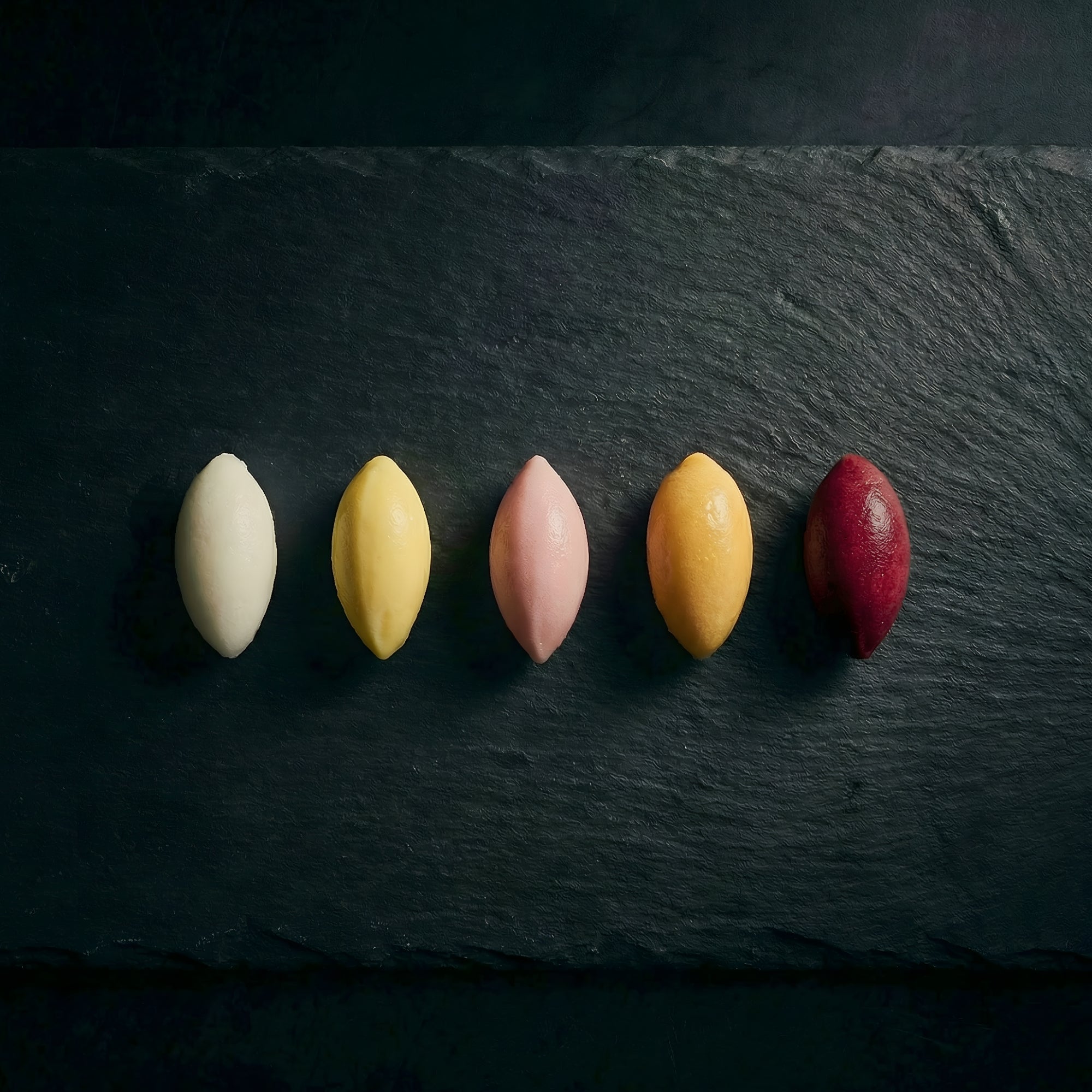 A row of vibrant, differently coloured ice cream rochers presented on a sleek black slate Michelin-style plate.