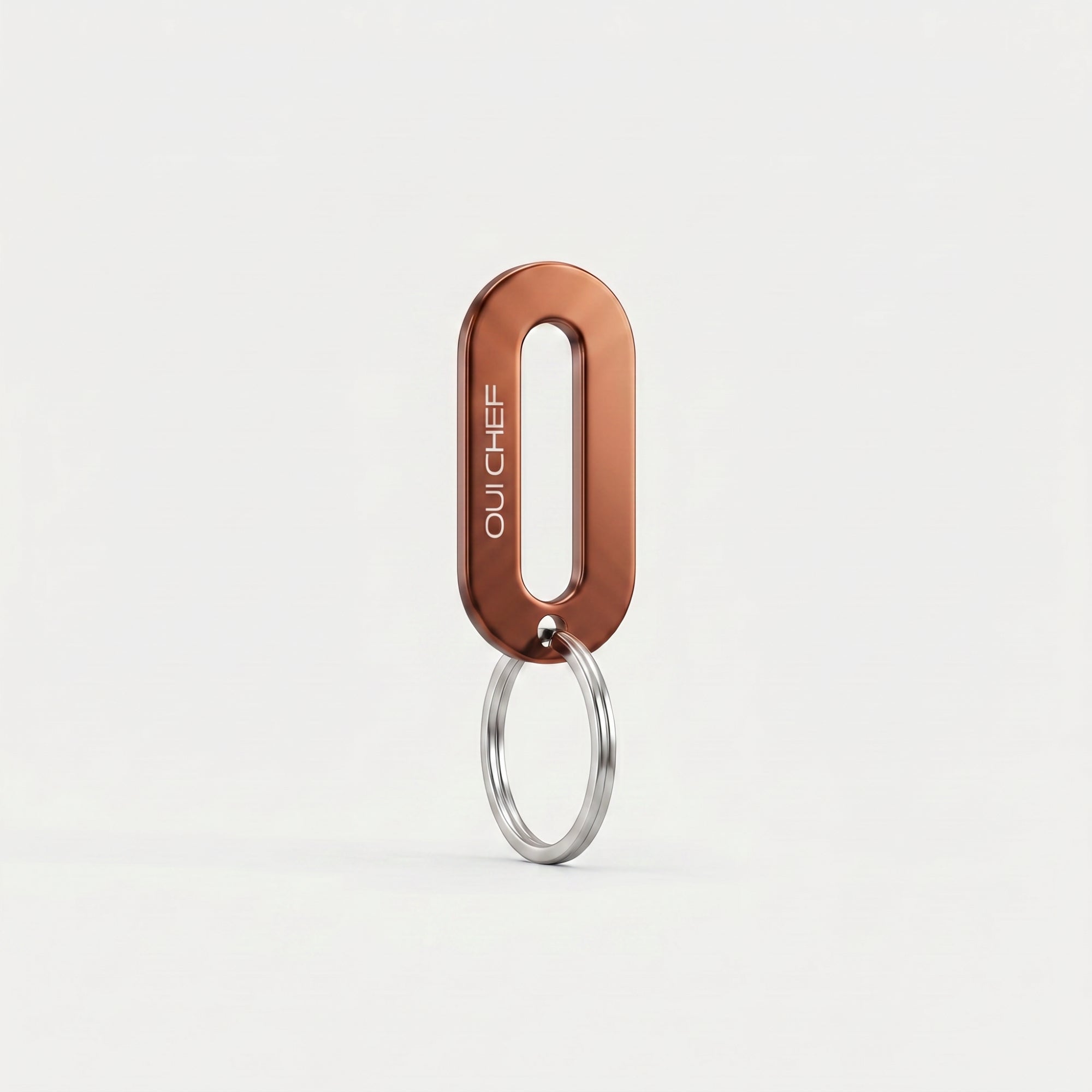 A copper colour keyring engraved with the Oui Chef logo styled standing upright on its ring in an artistic pose, set against a soft off-white background with subtle shadows highlighting its glossy finish.