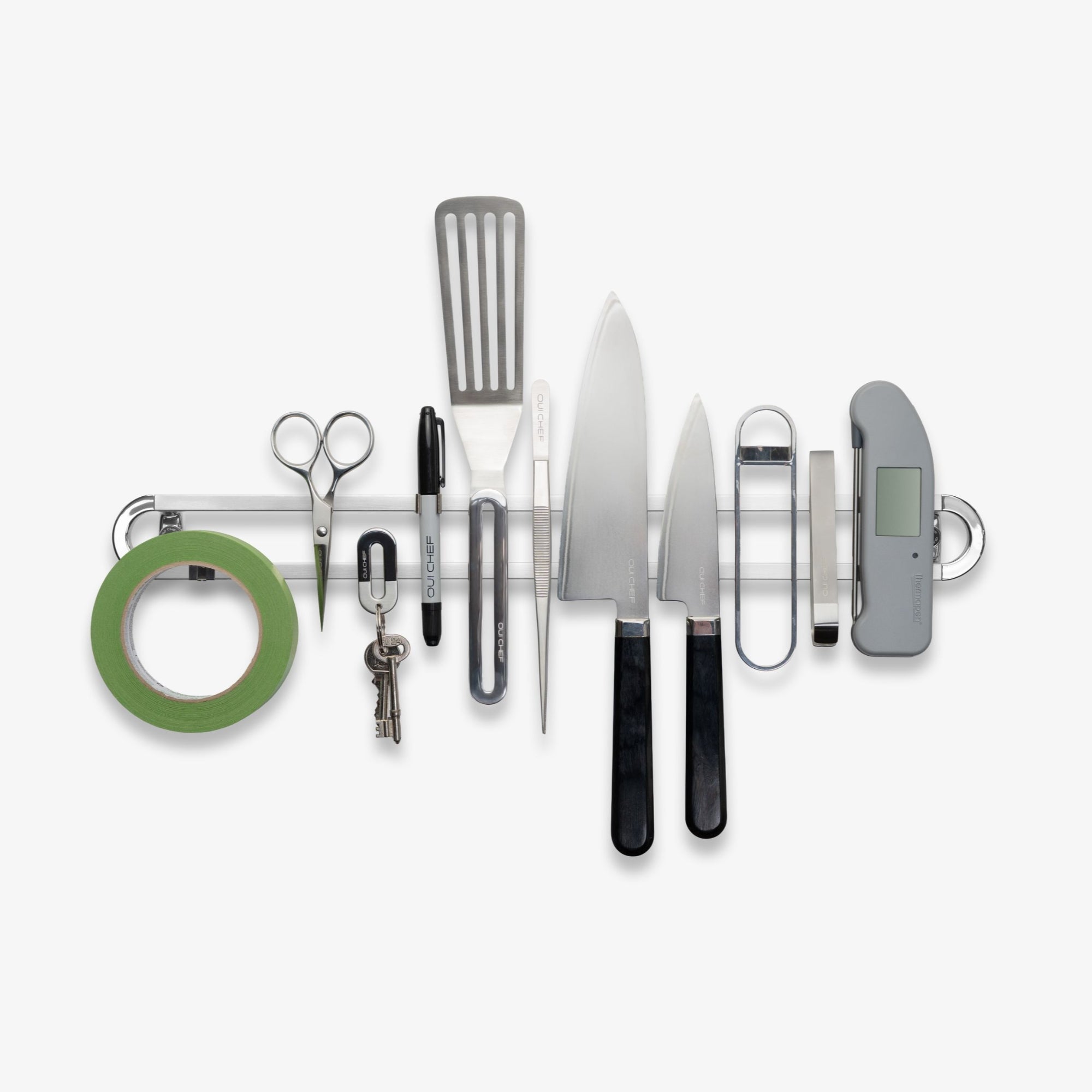 A Stainless steel magnetic rack sitting on a white background with the following line up from left to right; kitchen tape, scissors, keys, a sharpie, a spatula, tweezers, two chef's knives, a bottle opener, fish tweezers and a thermometer. 